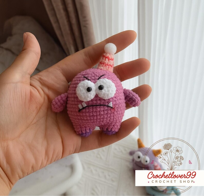 Pu&ograve; includere: Un giocattolo mostro fatto a mano, all'uncinetto in tonalit&agrave; di rosa e viola. Il giocattolo ha grandi occhi bianchi, un'espressione scontrosa e un cappello da festa. Il giocattolo &egrave; alto circa 7.6 cm.