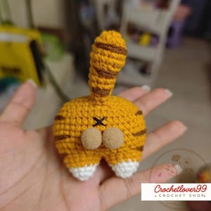 Op de afbeelding: Een handgemaakte gehaakte kattenkont in geel met bruine strepen en een staart. De kattenkont heeft twee beige pompons en een zwart geborduurd X. De onderkant van de kattenkont is wit. De afbeelding bevat de tekst "Crochetlover99 + CROCHET SHOP."