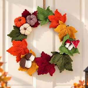 Peut inclure: Une couronne circulaire composée d'éléments d'automne crochetés, incluant des feuilles d'érable orange, jaunes, vertes et rouge foncé, des petites citrouilles orange, blanches et violettes, des glands marron et deux petits champignons rouges à pois blancs.