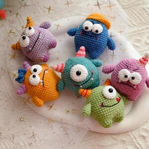 Pu&ograve; includere: Una collezione di sei mostri colorati all'uncinetto. I mostri sono blu, viola, arancioni e verdi, ognuno con grandi occhi a palla, corna e bocche sorridenti. Sono disposti su un piatto bianco, una collezione fatta a mano.