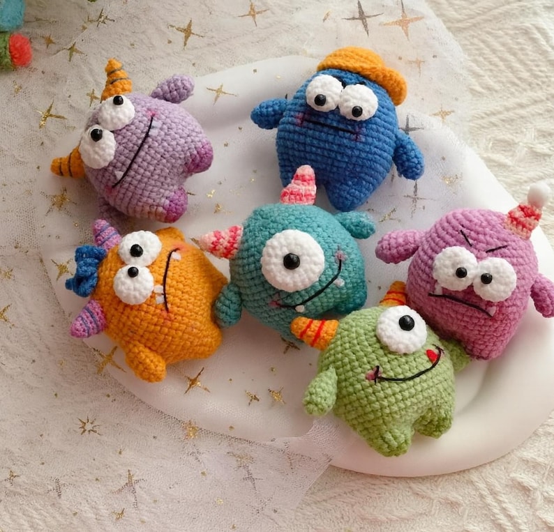 Pu&ograve; includere: Una collezione di sei mostri colorati all'uncinetto. I mostri sono blu, viola, arancioni e verdi, ognuno con grandi occhi a palla, corna e bocche sorridenti. Sono disposti su un piatto bianco, una collezione fatta a mano.