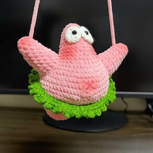 May include: A pink crocheted plush figure resembling a starfish with large white eyes, black pupils, and a green ruffled skirt around its waist. The plush is suspended by two pink yarn straps attached to its arms, featuring a soft, textured yarn.