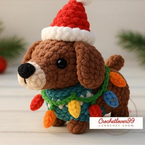 May include: A crocheted brown dog wearing a red and white Santa hat and a teal scarf adorned with colorful Christmas lights. The dog has a black nose and is a festive holiday decoration.