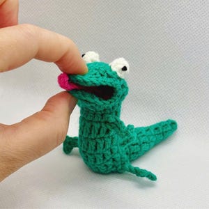 May include: A handmade green crochet alligator with white eyes and a pink tongue. The alligator is made of yarn and has a textured appearance. The alligator is a whimsical, handcrafted item.