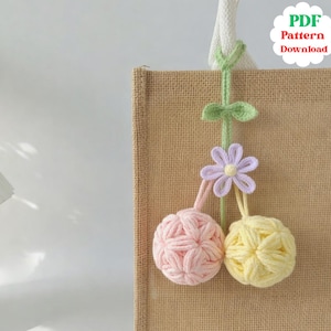 May include: A handmade crochet ornament with a floral design, featuring a light purple flower and two yarn ball ornaments in pink and yellow. The ornament is attached to a tan tote bag. Text on the upper right says "PDF Pattern Download".