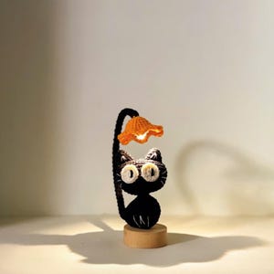 May include: A handmade black cat lamp featuring a curved stem and an orange flower lampshade. The cat has large white eyes and sits on a small wooden base. The lamp is made of yarn.