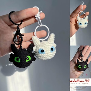 May include: Crocheted dragon keychains in black and white. The black dragon has green eyes, and the white dragon has blue eyes. Both have a round head, pointed ears, and are attached to a keychain ring with a clasp.