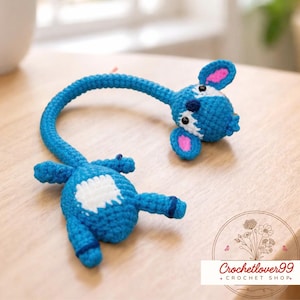 May include: A handmade blue crochet toy with a long, curved neck and a head with pink-lined ears. The toy has a white patch on its body and is lying on a wooden surface. The image also includes a logo that says "Crochetlover99 + CROCHET SHOP."