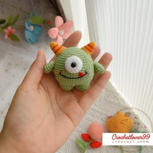 Pu&ograve; includere: Un mostro verde all'uncinetto fatto a mano con un occhio, corna arancioni e una lingua rossa. La piccola creatura rotonda ha una bocca sorridente e piccole braccia e gambe. Il mostro &egrave; tenuto in una mano, con uno sfondo sfocato di fiori.