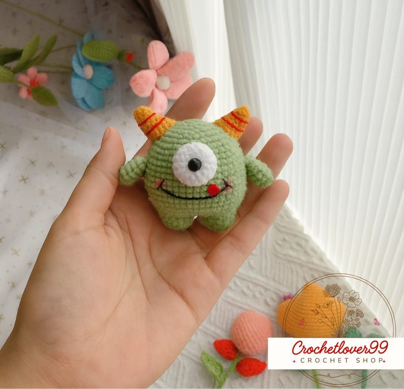 Pu&ograve; includere: Un mostro verde all'uncinetto fatto a mano con un occhio, corna arancioni e una lingua rossa. La piccola creatura rotonda ha una bocca sorridente e piccole braccia e gambe. Il mostro &egrave; tenuto in una mano, con uno sfondo sfocato di fiori.