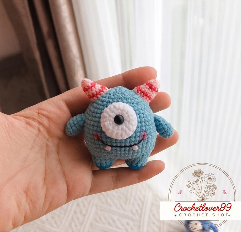 Pu&ograve; includere: Un piccolo giocattolo a forma di mostro all'uncinetto nei toni del blu, con un solo occhio bianco e corna a righe rosa e bianche. Il giocattolo ha braccia e gambe piccole e una bocca sorridente. Il testo "Crochetlover99 + CROCHET SHOP" &egrave; in basso a destra.