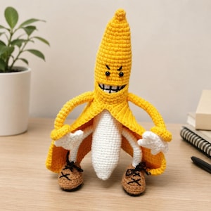 Könnte beinhalten: Eine gehäkelte Bananenfigur mit gelber Schale, weißem Fruchtfleisch und einem schelmischen Gesichtsausdruck. Die Figur hat dünne Arme, weiße Hände und braune Schuhe mit schwarzen Schnürsenkeln. Die Bananenschale ist offen und zeigt das Innere.
