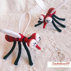 May include: Two handmade crochet mosquito toys. The mosquitoes are red and black with white wings and large white eyes. The legs are black. The toys are on a white, textured surface. The image includes a logo for a crochet shop.