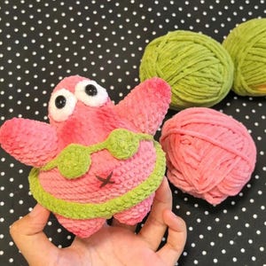 Può includere: Un peluche a forma di stella marina rosa con grandi occhi bianchi e neri, che indossa un bikini all'uncinetto verde chiaro. Il giocattolo è realizzato in filato morbido e testurizzato ed è posizionato accanto a due gomitoli di lana verde e uno rosa.
