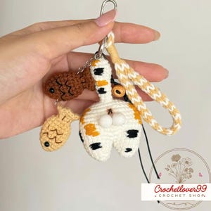 Op de afbeelding: Gehaakte kattenkont sleutelhanger met twee visbedels. De kattenkont is wit met oranje en zwarte vlekken. De vissen zijn bruin en beige. De sleutelhanger heeft een gevlochten gele en witte polsband.