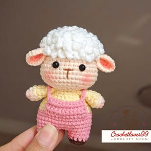 Op de afbeelding: Een handgemaakte gehaakte schapenfiguur met een pluizige witte kop, roze oren en roze wangen. Het schaap draagt een roze tuinbroek over een gele body. Zwarte knoopogen en een bruine 'T'-vormige mond maken het ontwerp compleet.