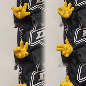 May include: Yellow crocheted hand keychains with various hand gestures, hanging on a black backpack. The keychains are attached to silver chains. The backpack has white lettering and a white border.