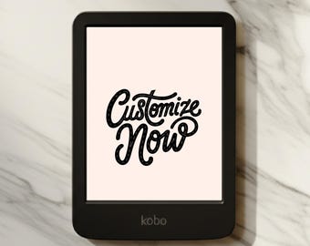 May include: A black Kobo e-reader with a light pink screen displaying the words "Customize Now" in black cursive script. The e-reader is on a white and gray marble surface.