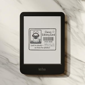 Peut inclure: Liseuse Kobo noire affichant une carte de bibliothèque avec le texte "Clara's Library Card" et "Lost in a book - no time for photos!". L'appareil est posé sur une surface en marbre.