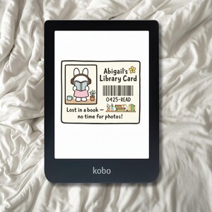 Könnte beinhalten: Ein schwarzer Kobo E-Reader mit einem Bibliothekskartendesign. Die Karte zeigt ein Cartoon-Kaninchen beim Lesen, den Text "Abigail's Library Card", einen Barcode und den Satz "Lost in a book - no time for photos!"