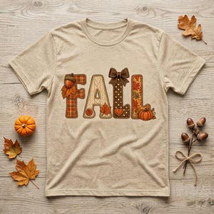 Könnte beinhalten: Ein kurzärmeliges T-Shirt in meliertem Beige mit dem Wort FALL in großen, dekorativen Buchstaben. Die Buchstaben sind mit Mustern aus Schottenkaro, Stricktexturen, Punkten und Herbstblättern gefüllt und mit einer Eichel, einer Schleife und einem Kürbis verziert.