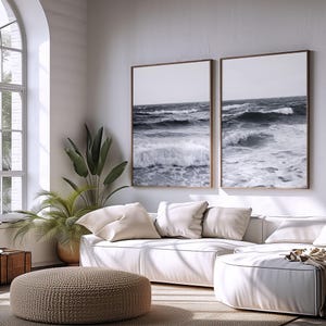 Può includere: Due stampe incorniciate in bianco e nero di onde oceaniche sono appese sopra un divano bianco. Le stampe hanno cornici marroni. Un pouf intrecciato e piante in vaso sono in primo piano. La stanza ha una palette di colori neutri.