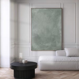 May include: A modern living room featuring a large, framed abstract painting in muted green tones. A black round coffee table with a ribbed base sits in front of a white sofa. Two candles in a gold holder and a small white bowl are on the table.