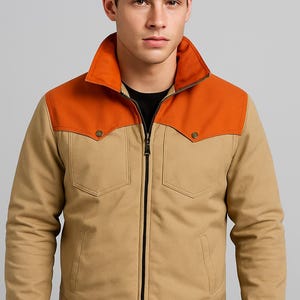 Op de afbeelding: Een beige en oranje jas met ritssluiting, twee voorzakken en een kraag. De jas heeft een contrasterende oranje pas op de schouders en is gemaakt van een duurzame stof.