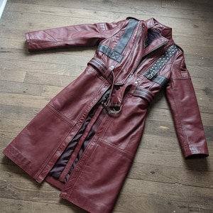 May include: A burgundy leather coat with black detailing, a belted waist, and studded embellishments. The long coat has a high collar and is displayed on a wooden surface. It features a zip closure and a dark interior lining.