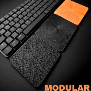 May include: A black computer keyboard positioned next to three square modular keyboard rests. One rest is bright orange with a sunburst pattern, while the other two are black with concentric circle and maze-like textures. Text reads MODULAR KEYBOARD REST.