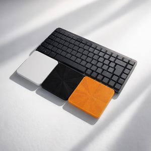 Könnte beinhalten: Eine schwarze Computertastatur, die über drei quadratischen Untersetzern platziert ist. Die Untersetzer sind in einer Reihe angeordnet und bestehen aus einem glatten weißen, einem schwarzen mit radialer Struktur und einem orangefarbenen mit gleichem Muster.
