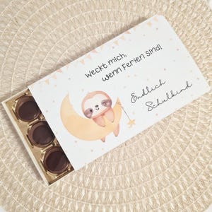 May include: A white gift box with a lid featuring a watercolor illustration of a sloth on a crescent moon, with text in German. The box contains six round chocolate candies. The box is on a beige woven placemat.