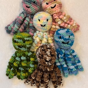 May include: A collection of seven handmade crochet octopuses in various colors. Each octopus has a round head with a smiling face and eight tentacles. The color palette includes pink, blue, green, and brown, with each octopus having a unique color combination.