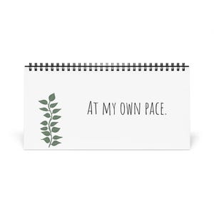 Peut inclure: Un calendrier de bureau blanc avec une reliure à spirale noire. Le calendrier affiche la phrase "AT MY OWN PACE" en noir, accompagnée d'une branche verte feuillue. Le calendrier est conçu pour être posé debout.