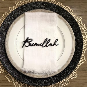 May include: A black and gold place setting with a white napkin and a black wooden place card that reads "Bismillah".