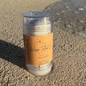 Puede incluir: Un tubo de plástico transparente con protector solar blanco en su interior. La etiqueta es marrón con el texto "Wisco Tallout Co." y "Sun Guard" en blanco. El tubo está sobre una playa de arena.