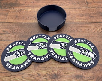 Könnte beinhalten: Set aus vier runden Untersetzern mit marineblauem Rand und den Worten "SEATTLE SEAHAWKS" um einen grünen Kreis mit einem weißen und grauen Falkenlogo. Ein marineblauer Untersetzerhalter ist ebenfalls enthalten.