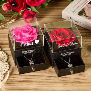 Puede incluir: Dos cajas de regalo negras con tapas transparentes, cada una con una rosa y un collar. Una caja tiene una rosa rosa y el nombre "Arthur" con un corazón. La otra tiene una rosa roja y los nombres "Garth & Olivia".