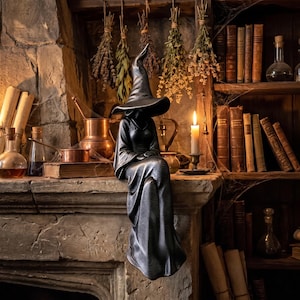 Peut inclure: Une figurine au ton métallique sombre représentant un personnage assis, vêtu d'une longue robe fluide et d'un chapeau de sorcière pointu. La silhouette est positionnée avec les jambes pendant au-dessus du bord d'une cheminée en pierre.
