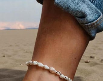 May include: Ankle bracelet made of small, white seashells strung together with black string. The bracelet is worn on an ankle, with a background of sand and blue jeans.