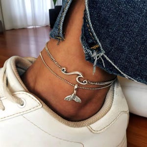 May include: Silver anklet set featuring a wave design and a small whale tail charm. The anklets are layered and worn on an ankle, paired with white sneakers and blue jeans.