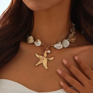 Może przedstawiać: Złoty naszyjnik z zawieszką w kształcie rozgwiazdy z perłowym akcentem. Naszyjnik posiada sznur nieregularnych, kremowo-białych i brązowych koralików. Zawieszka w kształcie rozgwiazdy jest teksturowana.