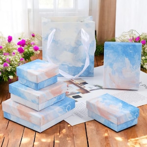 Puede incluir: Un conjunto de cuatro cajas de regalo azules y rosas con un diseño de nubes de acuarela. Las cajas están apiladas una encima de la otra y están rodeadas por una superficie blanca. También hay una bolsa de regalo azul y rosa con un diseño de nubes de acuarela.