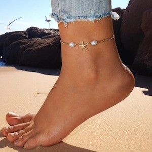 May include: A gold-coloured anklet with a starfish pendant and two faux pearls. The chain is worn around the ankle, with a light blue denim jean visible above. The anklet is a beach-themed jewellery item.