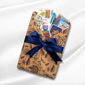 May include: A brown paper envelope with a black floral print tied with a dark blue satin ribbon. Inside are various stickers, a blue pen, and paper items. Visible text includes "BOOKS ARE MY LOVE" and "THERE'S A MILLION BOOKS".
