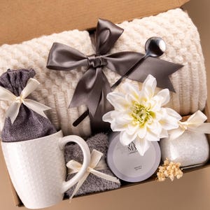 May include: A gift box containing a cream-colored textured throw blanket tied with a dark gray ribbon, a white ceramic mug with a dotted pattern, a gray fabric pouch, a gray candle, a glass jar with white bath salts, and a white dahlia flower.