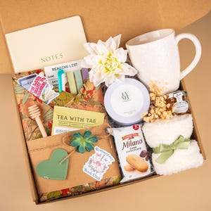 May include: A gift box filled with a white floral mug, a notepad, a candle, a jar of honey, a towel, and a package of Milano dark chocolate cookies. The box also contains a wooden honey dipper, stickers, and a decorative flower. The box is labeled "TEA WITH TAE" and "Romantasy Book Club."
