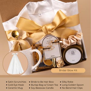May include: A gift box with a white silky robe, gold ribbon, and a ceramic mug. The box includes a gold eye mask, soy-beeswax candle, and hair accessories. The text "Bridal Glow Kit" is visible.