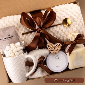 May include: A gift box containing a cream-colored textured throw blanket tied with a brown ribbon, a white mug with a scale pattern, a packet of marshmallows labeled Moonstruck, a gold spoon, a heart-shaped wicker ornament, and a candle.