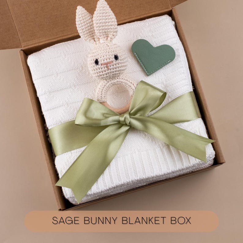 Puede incluir: Una manta blanca para beb&eacute; con textura de punto, una cinta verde salvia y un sonajero de conejito de ganchillo dentro de una caja de cart&oacute;n. Tambi&eacute;n se incluye una etiqueta en forma de coraz&oacute;n verde salvia. La caja tiene el texto "SAGE BUNNY BLANKET BOX".